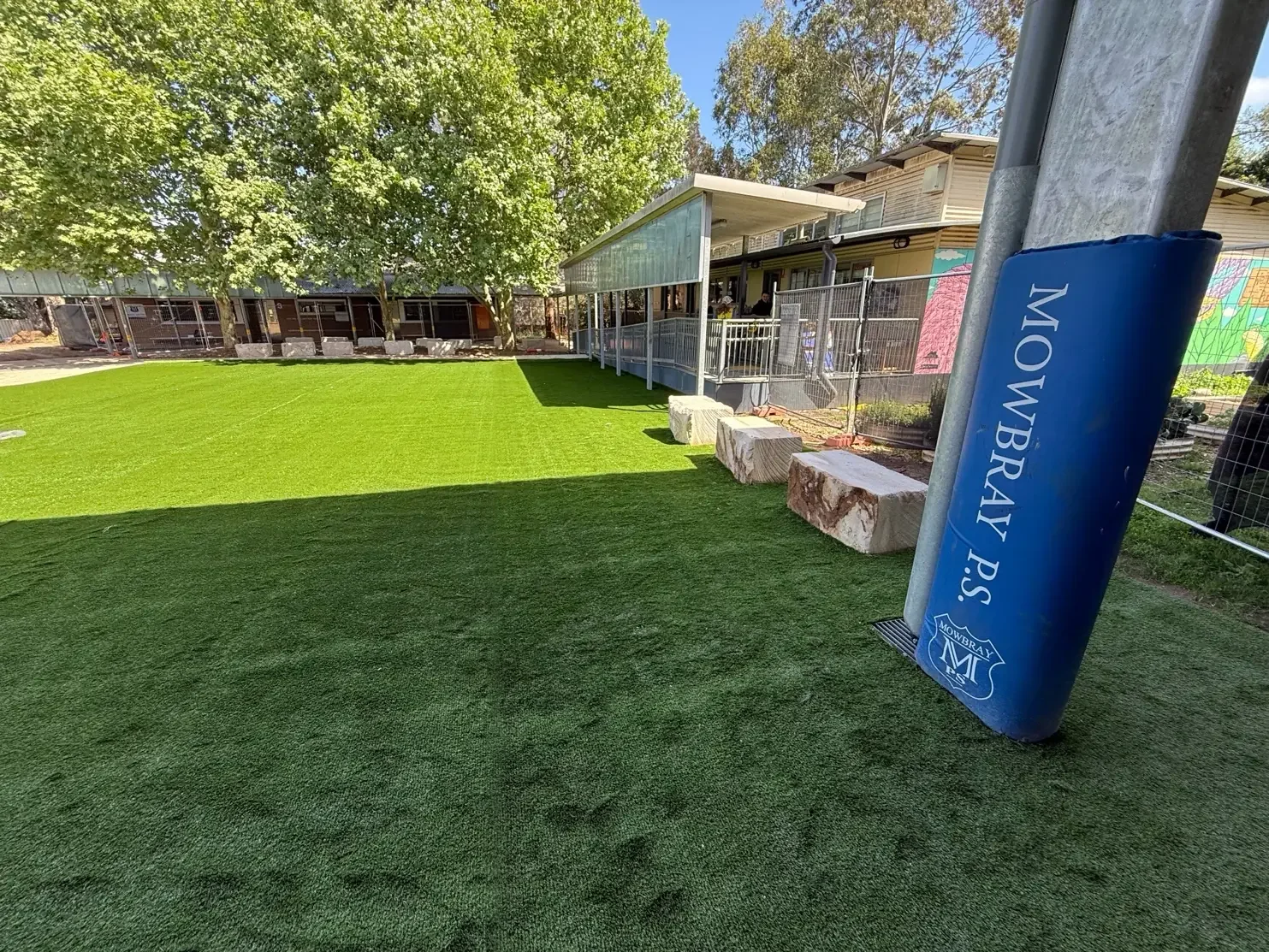 New synthetic grass beside a covered school walkway, with sandstone block seating and a padded goal post