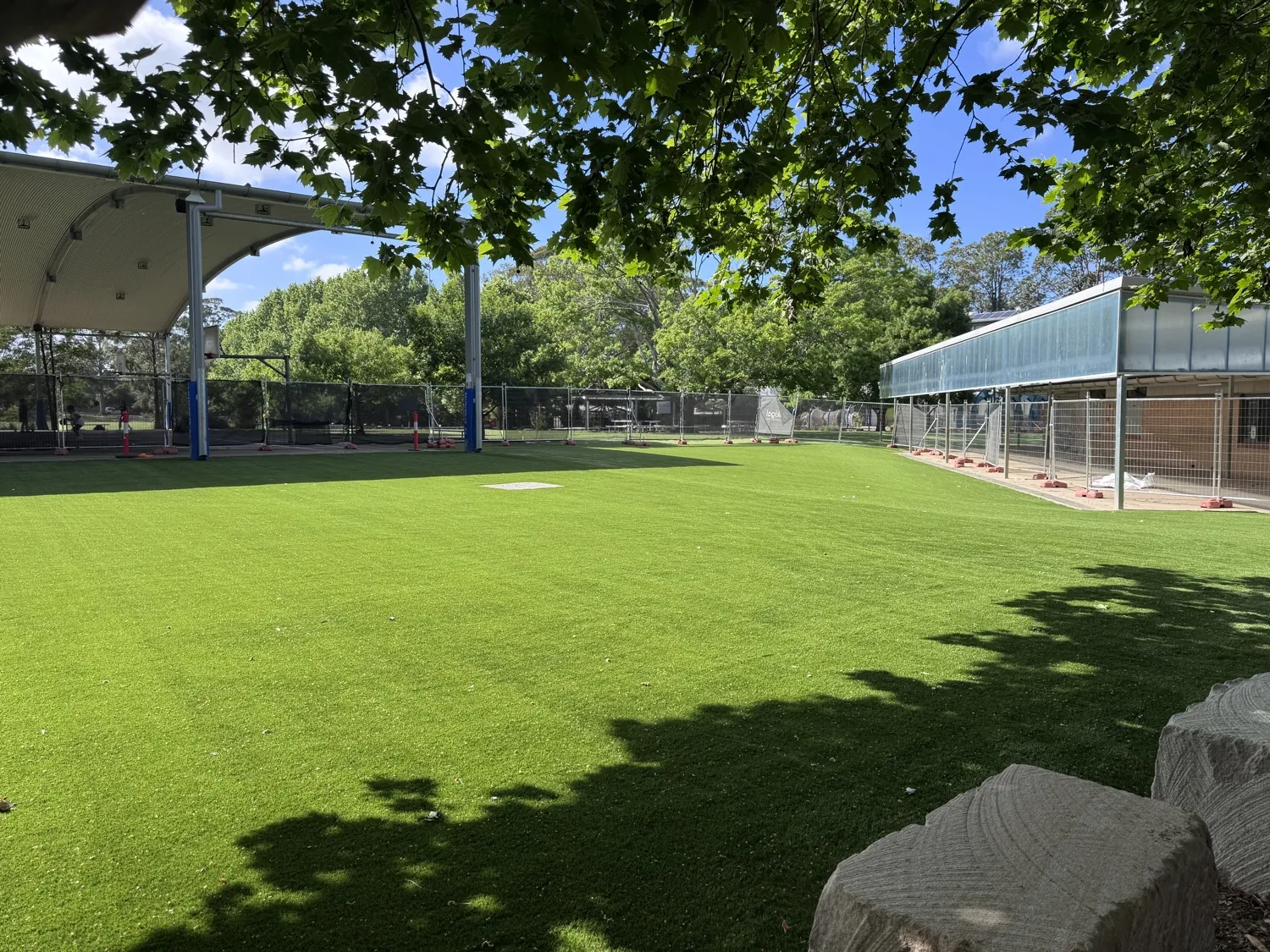 Synthetic grass field under the tree canopy, with the covered outdoor area behind and sandstone blocks in the foreground