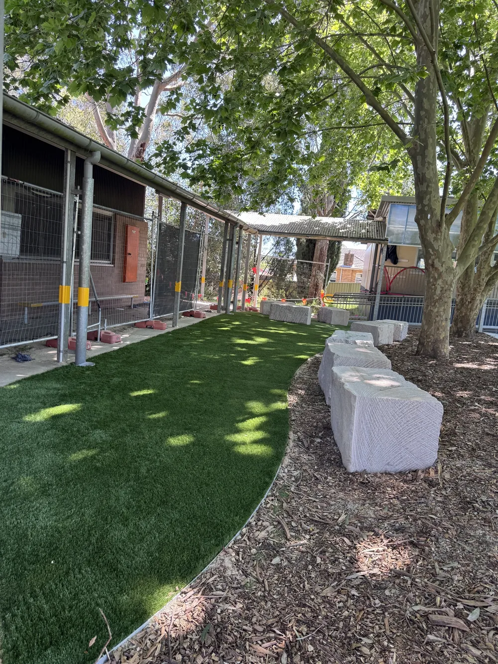 Curved synthetic grass edge along the covered walkway, with sandstone block seating in mulched garden beds under the trees